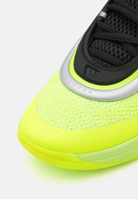 Neon-gelbe Sportschuh mit strukturiertem Mesh-Obermaterial, schwarzen Akzenten und einem silbernen Kunststoffdetail im Bereich der Schnürung. Glatter Gummisohle.