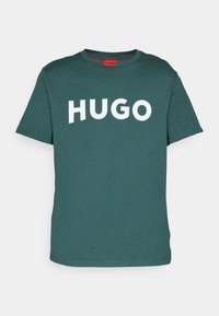 T-shirt en coton teal avec un col rond, des manches courtes et un logo blanc "HUGO" sur le devant. Texture lisse avec une coupe décontractée.