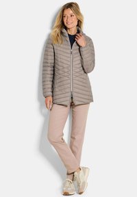 Vrouw staat, gekleed in een lichtbruine gewatteerde jas, beige broek en beige sneakers, kijkend opzij met een lichte glimlach.