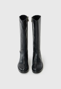 Bottes noires montantes en cuir lisse avec un bout arrondi, présentant un détail de fermeture éclair dorée sur le côté extérieur et une semelle plate.