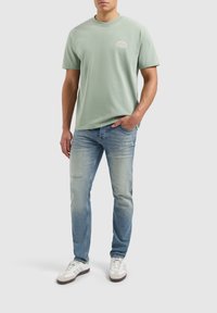 Lichtgroene katoenen t-shirt met korte mouwen en een ronde hals, gecombineerd met vervaagde blauwe jeans en witte sneakers met bruine accenten.