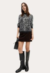 Strickpullover mit schwarz-weißem gepunktetem Muster, kombiniert mit einem braunen Cordrock, kniehohen schwarzen Lederstiefeln und einer schwarzen Handtasche.