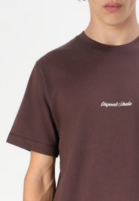 Ruda medvilninė marškinėliai trumpomis rankovėmis, su mažu baltu "Originals Studio" logo tekstu ant krūtinės. Lygi tekstūra.