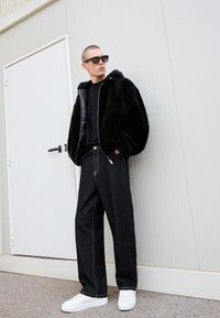 Uomo che indossa un giubbotto di pelliccia nera oversize, una maglietta nera, jeans scuri larghi, scarpe da ginnastica bianche e occhiali da sole neri, appoggiato a una parete bianca.