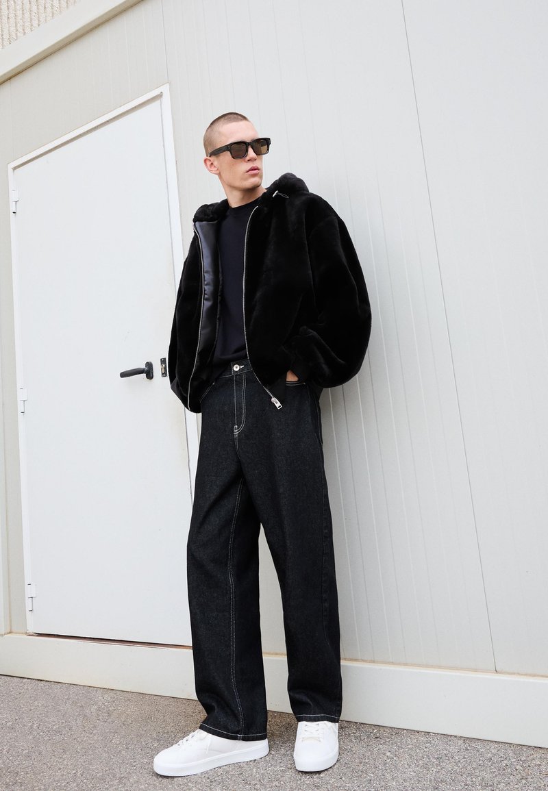 Uomo che indossa un giubbotto di pelliccia nera oversize, una maglietta nera, jeans scuri larghi, scarpe da ginnastica bianche e occhiali da sole neri, appoggiato a una parete bianca.
