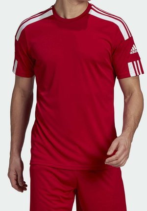 Man in een rood Adidas sportoutfit met witte strepen op de schouders en mouwen, staand met ontspannen armen.