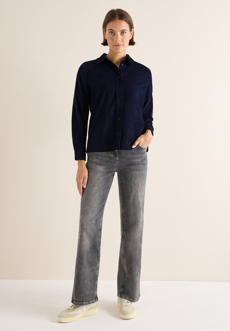 Camicia blu scuro con bottoni, realizzata in tessuto morbido e con maniche lunghe. Abbinata a jeans a gamba larga grigio chiaro e sneakers color crema. Design semplice, senza motivi visibili.