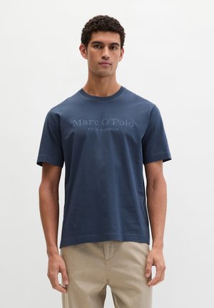 Mann mit kurzen lockigen Haaren, der ein blaues Marc O'Polo T-Shirt und beige Hosen trägt und vor einem schlichten weißen Hintergrund steht.