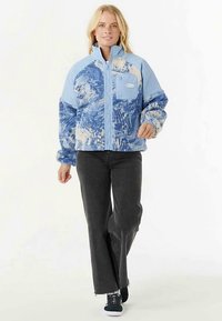 Blauwe fleecejas met abstract patroon in lichte en donkere blauwtinten, ritssluiting aan de voorkant, borstzak en hoge kraag. Draagt zwarte broek en sneakers.