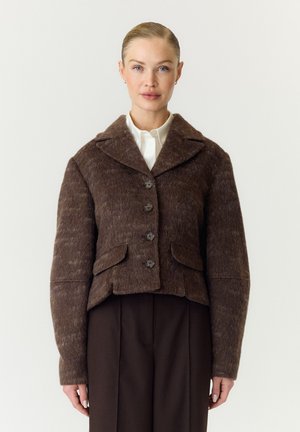 Frau mit blondem Haar, die eine braune, strukturierte, geknöpfte Jacke über einem weißen Hemd und dunkelbraunen Hosen vor einem schlichten Hintergrund trägt.