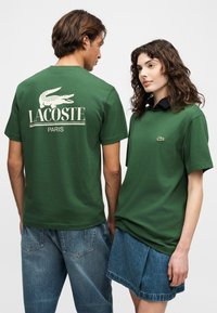 Άνδρας και γυναίκα που φορούν πράσινα t-shirts Lacoste· ο άνδρας είναι γυρισμένος προς τα πίσω και δείχνει μεγάλο λογότυπο, η γυναίκα κοιτάζει μπροστά, φοράει τζιν φούστα και μαύρο γιακά.