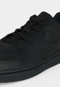 Primer plano de una zapatilla de cuero negra con cordones negros y el logo swoosh en relieve en el lateral sobre un fondo claro.