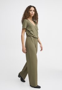 Combinaison vert olive à manches courtes, décolleté en V et taille élastique. Fabriquée en tissu doux, elle présente des jambes larges et droites ainsi que des chaussures noires.
