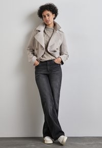 Cappotto doppiopetto in misto lana grigio chiaro, maglione beige a trecce, jeans neri a vita alta e sneakers bianche. Modella posa in modo casual.