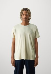 Camiseta de algodón verde claro con mangas cortas, que presenta un pequeño logo negro en el lado izquierdo del pecho. Textura suave, cuello redondo, corte relajado.