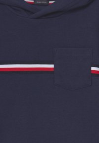 Holená navy modrá mikina z organické bavlny s ribbanou texturou, vybavená malou náprsní kapsou a horizontálním pruhem v červené, bílé a černé barvě.