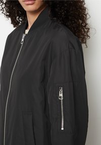 Mujer que lleva una chaqueta bomber negra con una cremallera plateada en el frente y un bolsillo con cremallera en la manga izquierda, su cabello rizado enmarca su rostro.