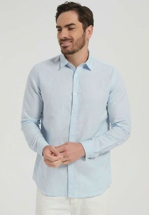 Homme aux cheveux bruns courts et à la barbe portant une chemise bleu clair boutonnée, debout et souriant, mains jointes.