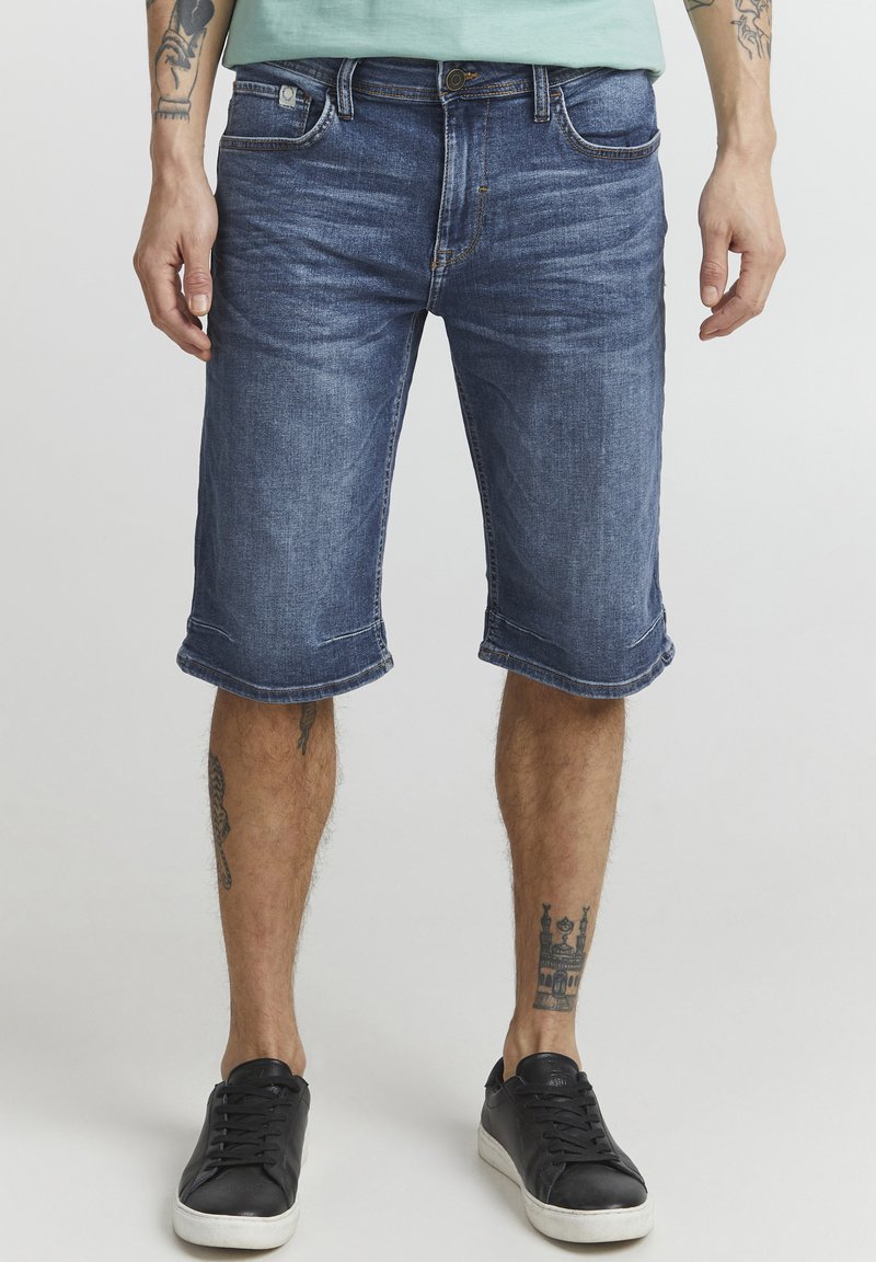 Pantalones cortos de mezclilla azul oscuro con dobladillos enrollados, diseño de cinco bolsillos y un sutil desvanecimiento. Combinados con zapatillas negras.