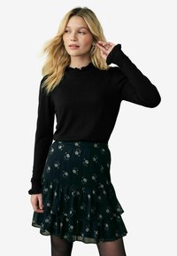 Top nero a maniche lunghe con polsini arricciati abbinato a una gonna a strati con motivo floreale verde scuro. Dettagli del tessuto testurizzato visibili.