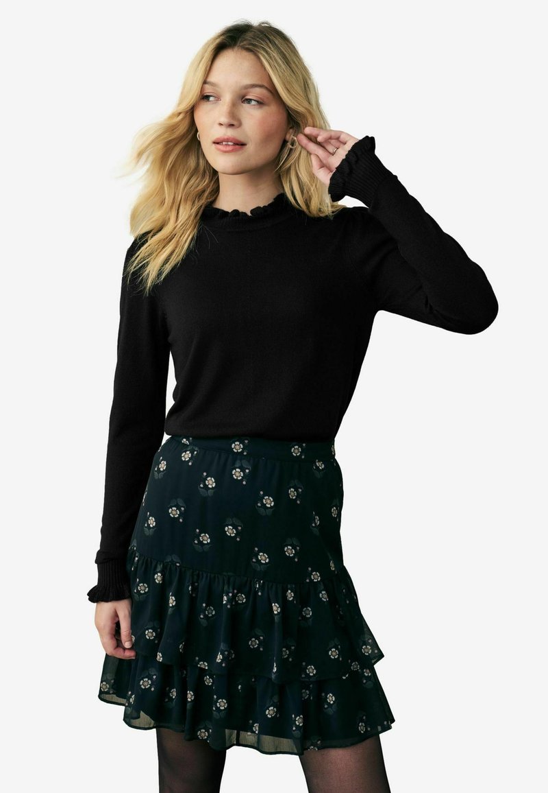 Top nero a maniche lunghe con polsini arricciati abbinato a una gonna a strati con motivo floreale verde scuro. Dettagli del tessuto testurizzato visibili.