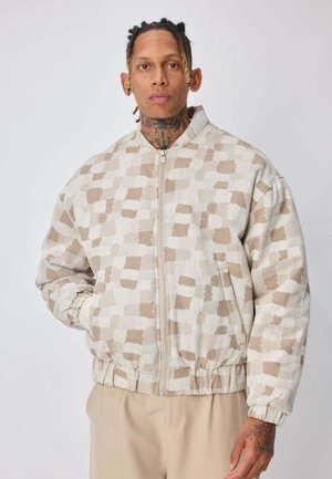 Homme aux cheveux tressés et tatouages sur le cou portant une veste zippée beige et blanche à carreaux et un pantalon beige, debout devant un fond uni.