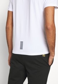 Camiseta blanca con un diseño minimalista, con rayas negras verticales en la parte posterior. Combinada con pantalones oscuros que tienen acentos dorados.
