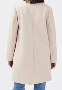 Beige frakke lavet af blødt stof, med et strømlinet design, lige snit, lange ærmer og en ren bagsøm. Båret over blå jeans.