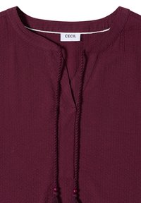 Chemise en tissu texturé violet foncé avec encolure fendue, cordons tressés se terminant par de petites perles et une étiquette blanche portant l'inscription « CECIL » à l'intérieur du col.