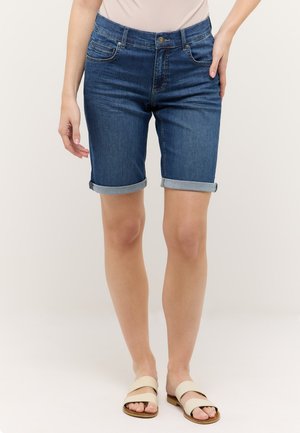 Mujer con pantalones cortos de mezclilla oscuros de tiro medio y sandalias beige sin talón, de pie contra un fondo liso.