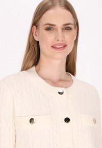 Kabelbrei crèmekleurige cardigan met een ronde hals, zilverkleurige knopen en voorzakken. Zachte textuur en gedetailleerd stikpatroon.