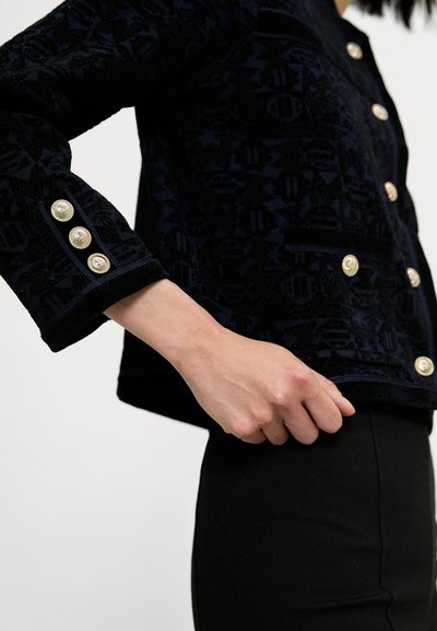Chaqueta de patrón navy con una tela texturizada, que presenta acentos de botones dorados en los puños y en la parte delantera, combinada con una parte inferior negra ajustada.