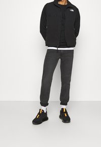 Svart fleeceväst med dragkedjor över en svart topp, i kombination med mörkgrå jeans och svarta sneakers med orange accenter. Vita strumpor synliga.