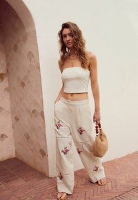 Top crema senza spalline abbinato a pantaloni beige chiaro a gamba larga con ricami floreali. Una borsa a tracolla intrecciata marrone e sandali semplici completano il look.