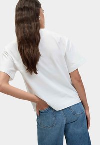 Vit kortärmad T-shirt med en avslappnad passform, parat med blå denimjeans. Håret är långt och vågigt, tucked bakom ett öra.