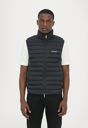 Zwarte puffer vest met hoge kraag, horizontale gestikte patronen, rits aan de voorkant en logo-detail op de linkerkant van de borst, gedragen over een wit shirt.