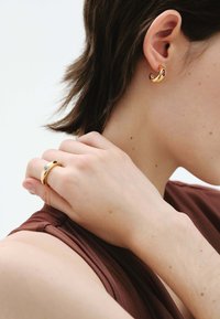 Pendientes de oro con un diseño curvado y acentos de piedras rojas. Un anillo de oro a juego con una piedra clara se ve en una mano cerca del cuello.