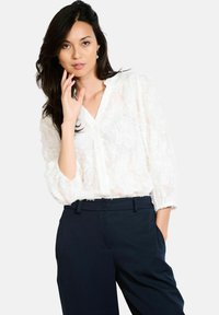 Blusa bianca e trasparente con texture floreale, chiusura a bottoni e scollatura a V. Abbinata a pantaloni blu scuro. Maniche lunghe arricciate ai polsini.