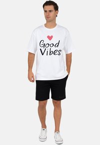 Wit T-shirt met korte mouwen met zwarte tekst "Good Vibes" en een rood hart. Gecombineerd met zwarte shorts en witte sneakers.