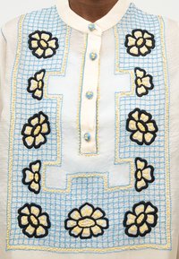 Λευκό πουκάμισο με μπλε καρό σχέδιο και κίτρινα κεντητά λουλούδια που περιβάλλουν το μπεζ πάπλωμα με κουμπιά και γιακά.