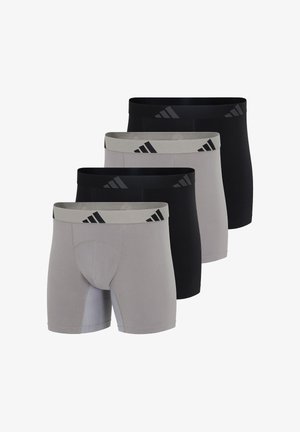 Atletyczny mężczyzna o ciemnej karnacji stojący boso, ubrany w jasnoszare bokserki Adidas z logo na gumie w pasie, patrzący nieco na bok.