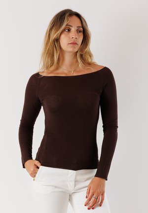 Top lungo marrone con maniche a lungo, scollatura ampia e design aderente. Realizzato in materiale morbido ed elastico, abbinato a pantaloni bianchi.