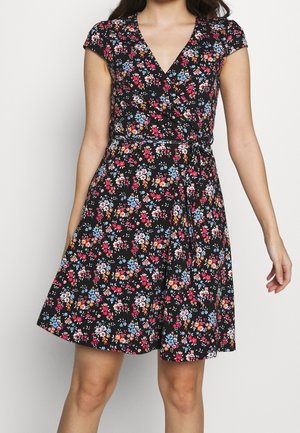 Frau trägt ein schwarzes Wickelkleid mit Blumenmuster in Rot, Blau und Weiß, kurzen Ärmeln und V-Ausschnitt.