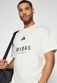 Biela tričko s okrúhlym výstrihom a krátkymi rukávmi. Obsahuje čierne logo "DIDAS SPORTSWEAR" a tri čierne šikmé pruhy. Materiál: bavlna.