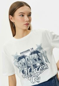 Camiseta blanca de algodón con un estampado gráfico que presenta "Brooklyn" y "New York City" en texto gris desgastado, acentuado por estrellas azules.