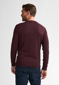 Burgunderfarbener Strickpullover mit Rundhalsausschnitt, langen Ärmeln und gerippten Bündchen. Getragen mit dunklen Hosen, die einen strukturierten Stoff zeigen.