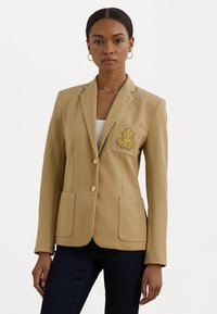 Blazer beige ajusté avec deux boutons, poche simple ornée des initiales jaunes brodées et deux poches avant. Tissu à texture lisse.