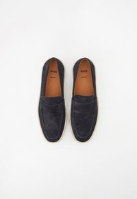 Navy suede loafers dengan desain moccasin yang dijahit, ujung bulat, dan sol karet berwarna terang yang kontras. Dilengkapi dengan bagian atas yang empuk.