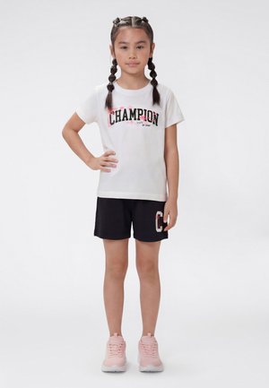 Mädchen mit geflochtenem Haar, trägt ein weißes "Champion"-T-Shirt, schwarze Shorts mit Logo und pinke Turnschuhe, steht mit einer Hand in der Hüfte.