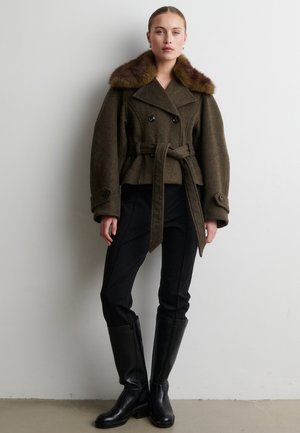 Donna che indossa un cappotto marrone con cintura e collo in pelliccia, pantaloni neri aderenti e stivali neri al ginocchio, in piedi contro un muro bianco semplice.